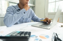 Άτομο μπροστά από λάπτοπ με χαρτιά που απεικονίζουν γραφήματα κάνει υπολογισμούς