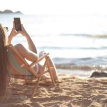 Γυναίκα σε ξύλινη καρέκλα στην παραλία κοιτά το κινητό της τηλέφωνο