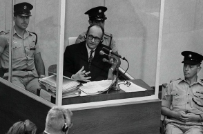 Ο Ναζί εγκληματίας πολέμου Άντολφ Άιχμαν στέκεται σε προστατευτικό γυάλινο θάλαμο πλαισιωμένο από την ισραηλινή αστυνομία κατά τη διάρκεια της δίκης του στις 22 Ιουνίου 1961 στην Ιερουσαλήμ