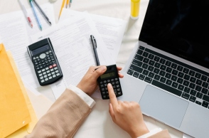 Δύο χέρια χειρίζονται calculator πάνω από ένα laptop μαζί με λογιστικά έγγραφα