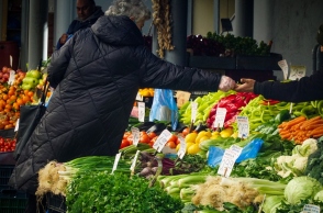 Γυναίκα όρθια με μαύρο παλτό δίνει λεφτά σε άνδρα με μαύρο σκούφο πάνω από τελάρα με λαχανικά
