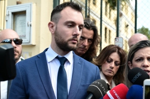 Άνδρας με κοστούμι μπροστά σε μικρόφωνα ανάμεσα από δημοσιογράφους