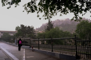 Άνθρωπος στα μαύρα περπατά σε δρόμο δίπλα σε κάγκελα με κόκκινη τσάντα