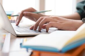 Γυναικείο χέρι πληκτρολογεί σε laptop και δίπλα υπάρχει ένα βιβλίο