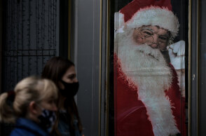 Πολίτες με μάσκα για τον κορωνοϊό στη Θεσσαλονίκη με φόντο εικόνα του Άγιου Βασίλη