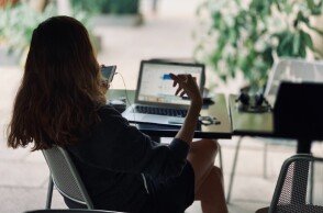 Κοπέλα κάθεται στο γραφείο της με ανοιχτό λάπτοπ και κρατάει το κινητό της τηλέφωνο©Unsplash/ hj barraza