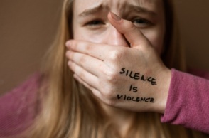 Μια γυναίκα που κλαίει, κλείνει το στόμα της και το χέρι της γράφει «η σιωπή είναι βία».