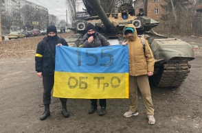 Τρεις νεαροί άνδρες κρατούν σημαία της Ουκρανίας μπροστά από τανκ