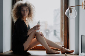 Κοπέλα με σγουρά μαλλιά καθισμένη δίπλα σε παράθυρο φορώντας μαύρα ρούχα βάζει κρέμα στα πόδια της