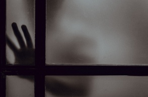 Χέρι και πρόσωπο διακρίνεται πίσω από κλειστή πόρτα σε ημίφως