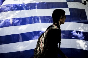 Γυναίκα προχωρά μπροστά από ελληνική σημαία που κυματίζει