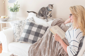 Γυναίκα καθισμένη σε καναπέ διαβάζει βιβλίο, δίπλα κάθεται γατάκι