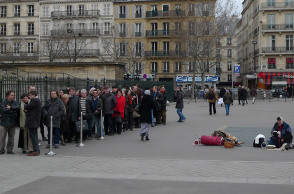 Άνθρωποι που περιμένουν σε μια ουρά και μια άστεγη γυναίκα κάθεται κοντά τους στην πλατεία Madeleine στο Παρίσι