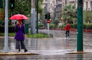 Γυναίκα με ομπρέλα περιμένει σε διάβαση πεζών την ώρα που ποδηλάτης με αδιάβροχο κινείται στους δρόμους της Αθήνας