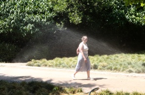 Γυναίκα περπατά σε συνθήκες καύσωνα σε περιβάλλον όπου ποτιστικό δροσίζει φυτά πετώντας νερό