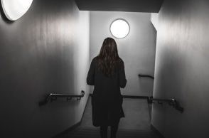 Γυναίκα περπατά με την πλάτη γυρισμένη στον φακό προς πόρτα που βρίσκεται σε κατηφορικό σκοτεινό διάδρομο