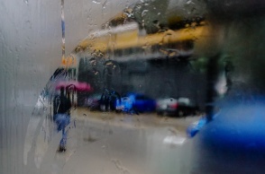 Φωτογραφία μέσα από αυτοκίνητο με θολωμένα τζάμια που δείχνει γυναίκα να κρατά ομπρέλα κοντά σε συρμό του τραμ που διασχίζει τον βροχερό δρόμο