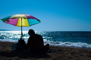 Άνδρας καθισμένος κάτω από πολύχρωμη ομπρέλα θαλάσσης στην ακροθαλασσιά