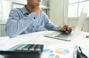 Άτομο μπροστά από λάπτοπ με χαρτιά που απεικονίζουν γραφήματα κάνει υπολογισμούς