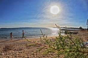 Ακτίνες του ήλιου πέφτουν στον φωτογραφικό φακό ενώ τραβά σκηνή από παραλία με αμμουδιά και δύο άτομα με μαγιό να βρίσκονται στην ακρογυαλιά