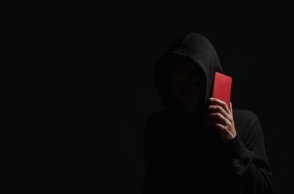 Άτομο με κουκούλα σε σκοτεινό δωμάτιο κρατά κόκκινο κινητό