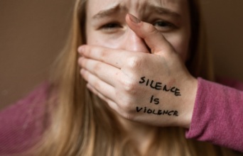 Μια γυναίκα που κλαίει, κλείνει το στόμα της και το χέρι της γράφει «η σιωπή είναι βία».