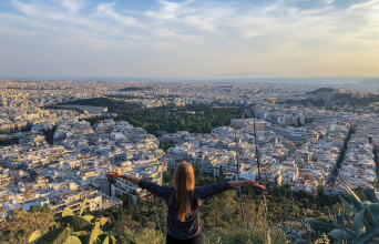 Κοπέλα στέκεται όρθια με γυρισμένη πλάτη με φόντο την Αθήνα από ψηλά