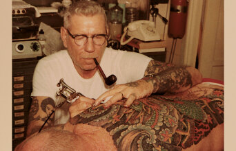 Norman Collins tattooing back with pipe in mouth