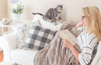 Γυναίκα καθισμένη σε καναπέ διαβάζει βιβλίο, δίπλα κάθεται γατάκι