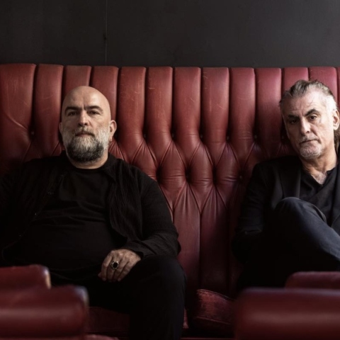 Ο Φίλιππος Πλιάτσικας και ο Μπάμπης Στόκας καθισμένοι σε κόκκινο καναμπέ φορώντας μαύρα ρούχα, κοιτούν το φακό