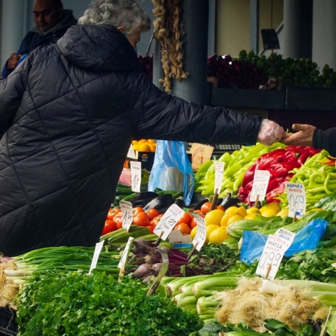 Γυναίκα όρθια με μαύρο παλτό δίνει λεφτά σε άνδρα με μαύρο σκούφο πάνω από τελάρα με λαχανικά