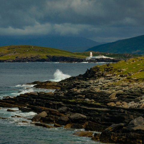 Βραχώδης παραλία και στο βάθος άσπρο κτίριο και κόκκινος φάρος