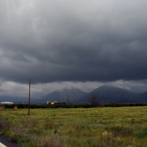 Επαρχιακός δρόμος δίπλα σε πράσινο χωράφι - Στο βάθος βουνά και σύννεφα