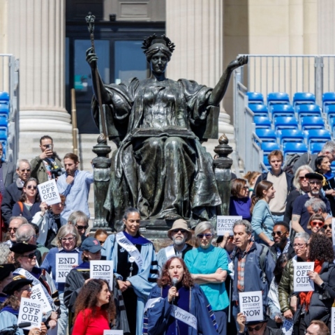 Καθηγητές ντυμένοι με παραδοσιακές ρόμπες μπροστά από άγαλμα κρατούν πλακάτ