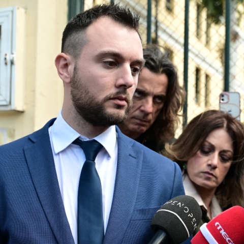 Άνδρας με κοστούμι μπροστά σε μικρόφωνα ανάμεσα από δημοσιογράφους