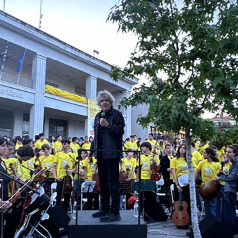 Άνδρας με μικρόφωνο πάνω σε βάθρο, ανάμεσα από παιδιά στα κίτρινα με μουσικά όργανα