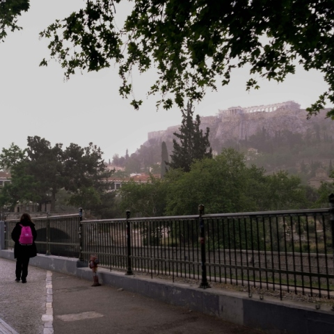 Άνθρωπος στα μαύρα περπατά σε δρόμο δίπλα σε κάγκελα με κόκκινη τσάντα