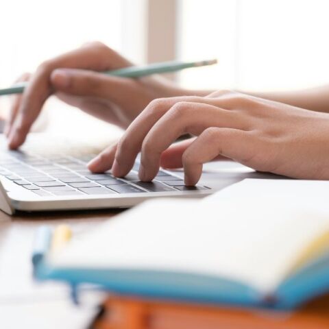 Γυναικείο χέρι πληκτρολογεί σε laptop και δίπλα υπάρχει ένα βιβλίο