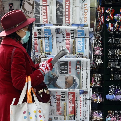 Γυναίκα που φορά ιατρική μάσκα προστασίας στέκεται μπροστά σε περίπτερο με γαλλικές εφημερίδες