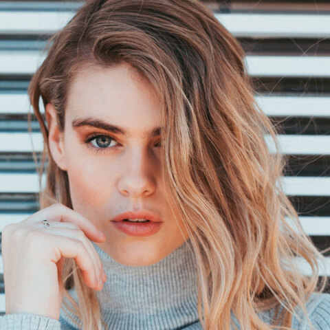 Ξανθιά κοπέλα με γκρι πλεκτό ζιβάγκο και φόντο ξύλινο φράκτη