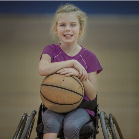 Κοριτσάκι καθισμένο σε αναπηρικό καρότσι χαμογελάει κρατώντας μια μπάλα μπάσκετ στα χέρια της