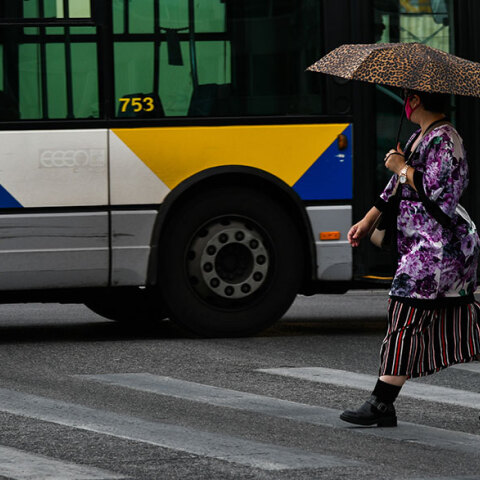Γυναίκα διασχίζει διάβαση πεζών στον Πειραιά, κρατώντας ομπρέλα και φορώντας προστατευτική μάσκα για τον κορωνοϊό