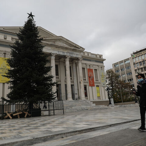 Γυναίκα με προστατευτική μάσκα περνά μπροστά από το Δημοτικό Θέατρο Πειραιά, όπου έχει στηθεί ένα τεράστιο χριστουγεννιάτικο δέντρο