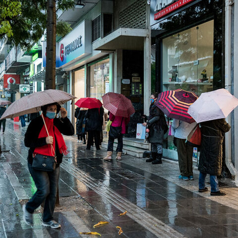 Πολίτες με ομπρέλες και μάσκες για τον κορωνοϊό σε πεζόδρομο εν μέσω βροχής