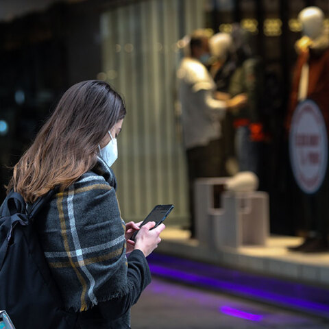 Κοπέλα με μάσκα για τον κορωνοϊό κοιτάζει το κινητό της με φόντο καταστήματα στην Ερμού