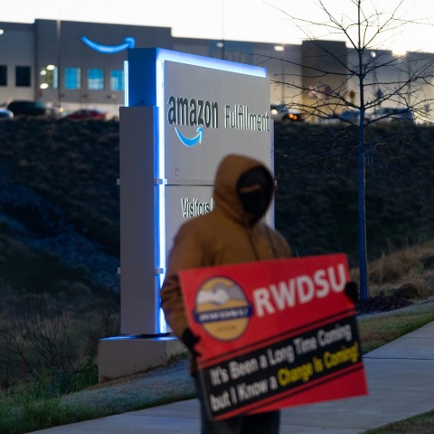 Εργαζόμενος κρατά πλακάτ έξω από αποθήκη της Amazon