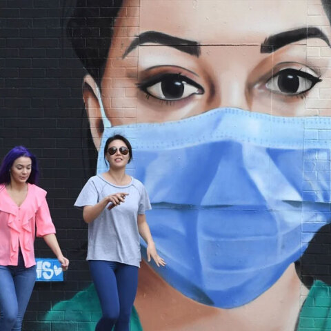 Νεαρές κοπέλες χωρίς μάσκα περπατούν στο Λονδίνο με φόντο ένα γκράφιτι για την πανδημία Covid-19