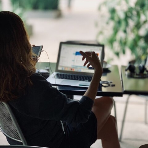 Κοπέλα κάθεται στο γραφείο της με ανοιχτό λάπτοπ και κρατάει το κινητό της τηλέφωνο©Unsplash/ hj barraza