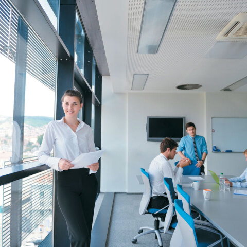 Κοπέλα όρθια σε χώρο γραφείου, στο βάθος άντρες καθιστοί γύρω από τραπέζι