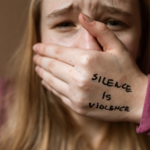Μια γυναίκα που κλαίει, κλείνει το στόμα της και το χέρι της γράφει «η σιωπή είναι βία».
